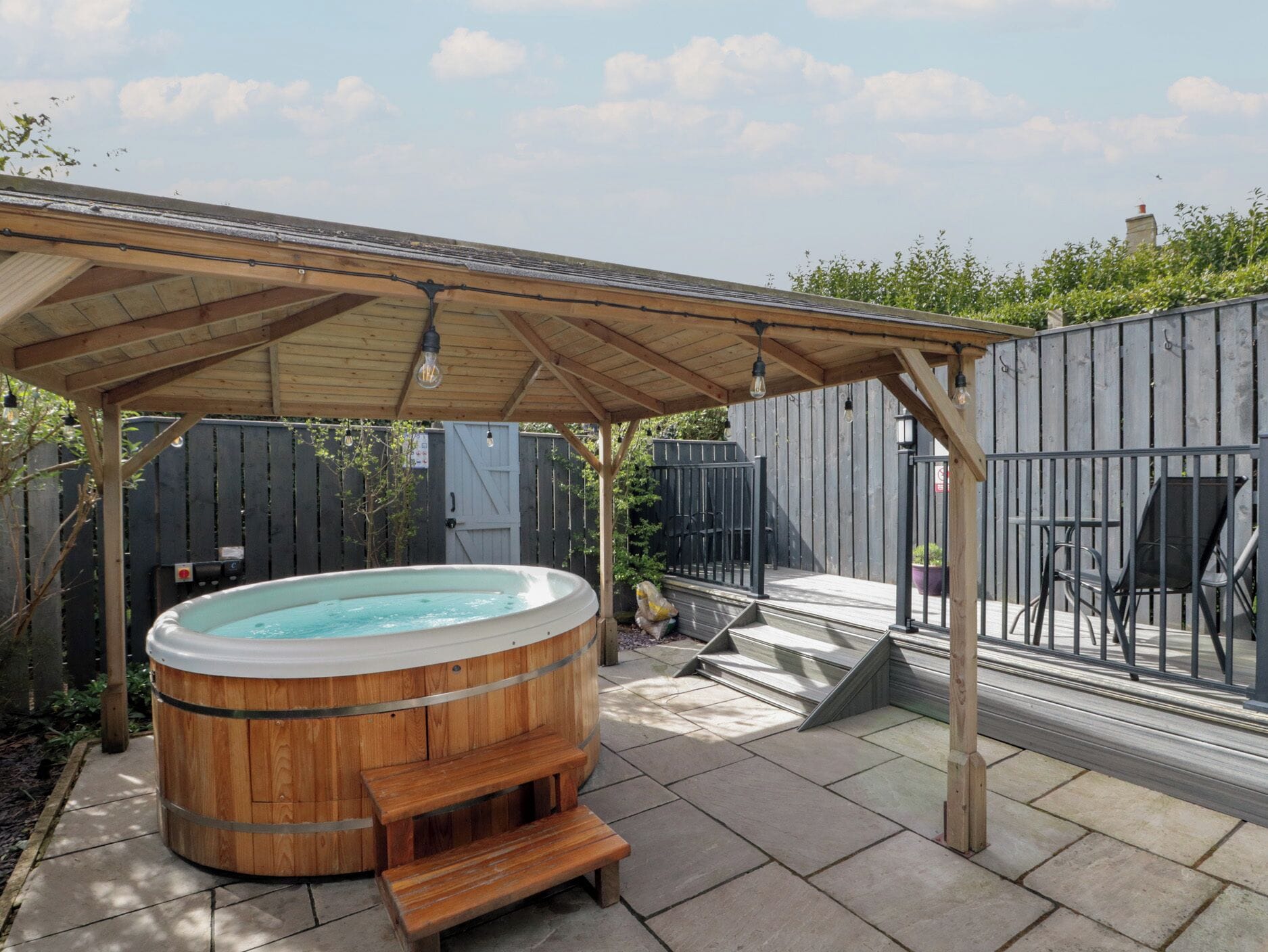 A wooden hot tub under a pergola with steps leading up to it, surrounded by a patio area with chairs and a fence.