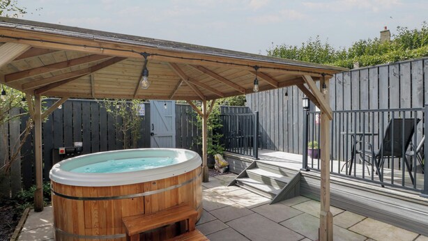 A wooden hot tub under a pergola with steps leading up to it, surrounded by a patio area with chairs and a fence. - Isla Cottage (Northallerton)