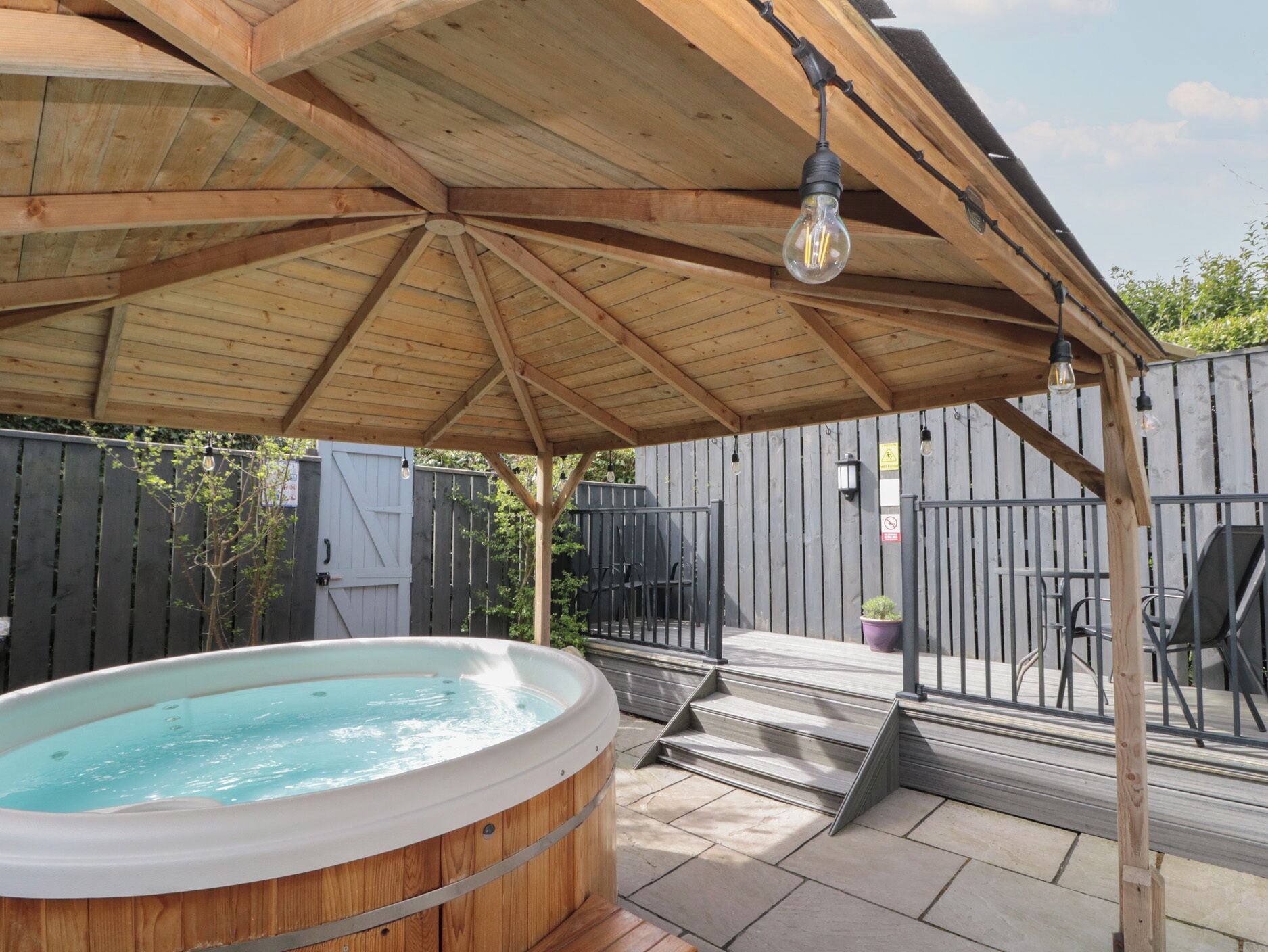 A wooden deck with a hot tub, a pergola with hanging lights, and a fenced backyard.