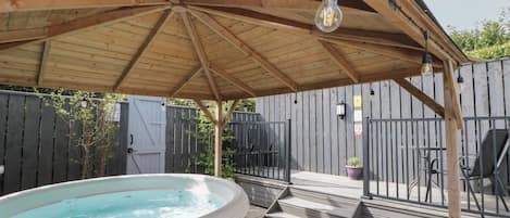 A wooden deck with a hot tub, a pergola with hanging lights, and a fenced backyard.