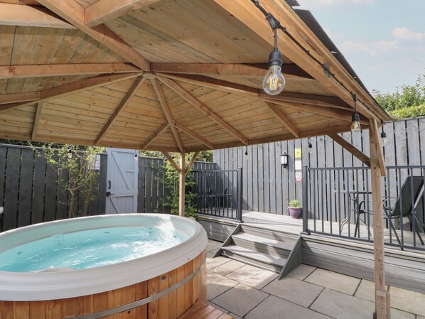 Una terraza de madera con un jacuzzi, una pérgola con luces colgantes y un patio trasero cercado.