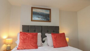 A bed with a grey upholstered headboard, white bedding, and two red pillows. There is a framed picture above the bed, and two bedside lamps on either side. - Isla Cottage (Northallerton)