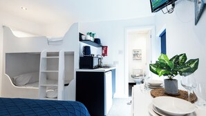 Een compacte kamer met een stapelbed, een kitchenette, een eethoek met een tafel voor twee en een potplant.