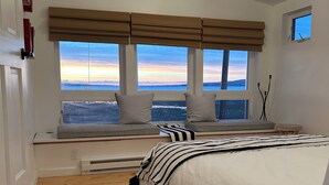 Un dormitorio con una cama grande, un rincón con ventanilla y cojines, y vistas a una puesta de sol sobre el agua.