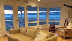 Un salón espacioso con un amplio sofá seccional color beige, suelo de madera y vistas a un lago a través de amplias ventanas.
