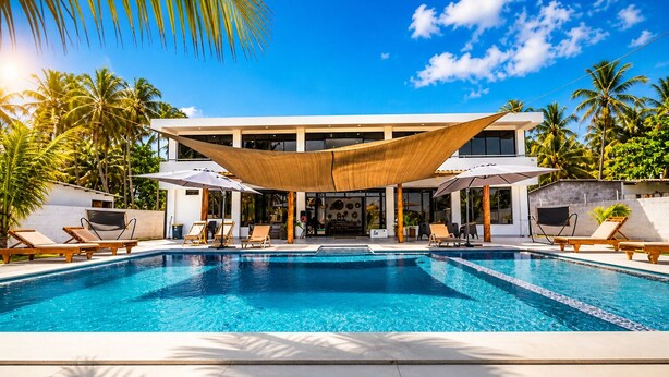 Un área de piscina con sillones de playa y sombrillas, un edificio de diseño moderno, y palmeras al fondo.