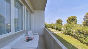 A balcony with a white table and chair, a magazine, and a view of a grassy area with trees and a distant building. - Prime Coastal Location Right in Front of the sea (Lignano Sabbiadoro)