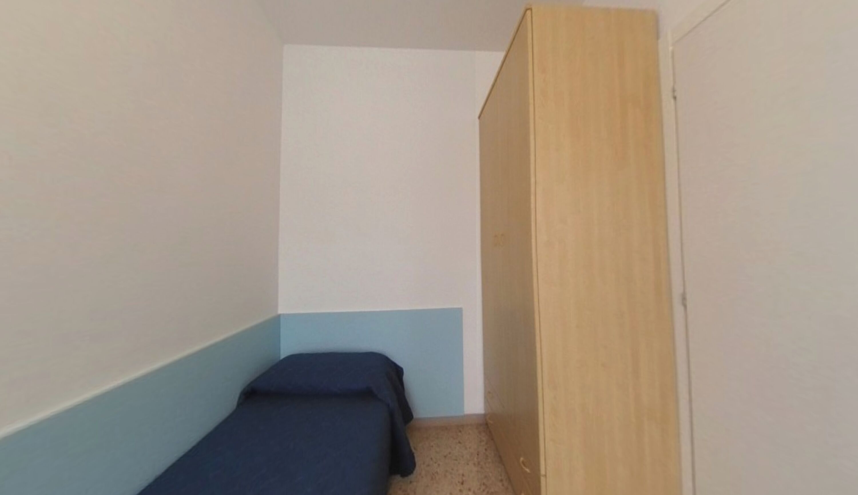 A small, narrow room with a single bed, a wooden wardrobe, and a light blue wall.