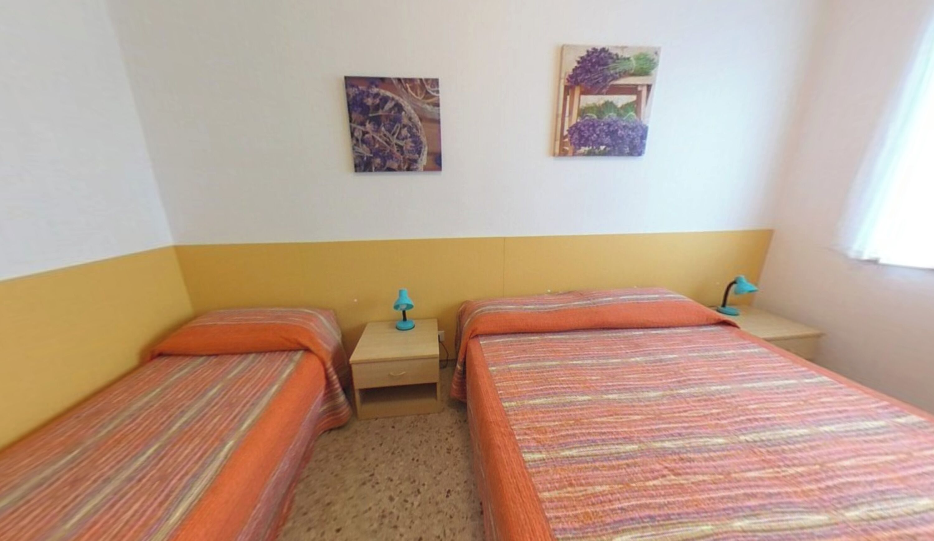 A room with two beds, a nightstand, a lamp, and two wall-mounted pictures.