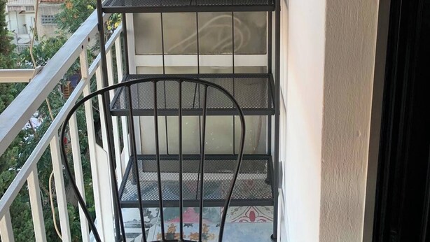 Un balcón con una repisa metálica que sostiene macetas y una silla.