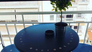 Mesa redonda con dos sillas, una planta en maceta y un posavasos pequeño en un balcón con baldosas estampadas y barandilla.