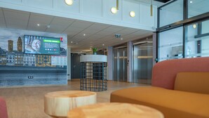 Un hall d’entrée moderne doté d’un grand écran numérique affichant une vue sur la ville, d’un comptoir d’accueil, d’une plante et d’un espace détente avec des tabourets en bois.