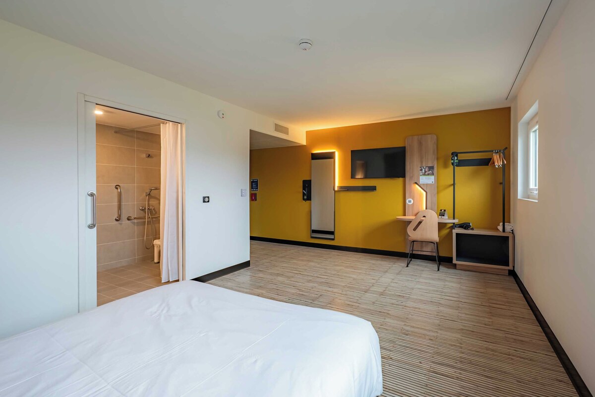 Habitación de hotel con cama, escritorio, silla, televisor y zona de ducha.