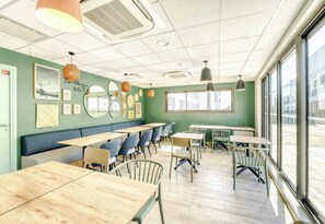 Un café con mesas y sillas de madera, una pared verde con obras de arte y ventanas grandes.