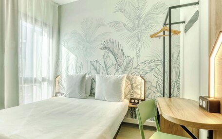 A hotel room with a bed, a desk, a chair, a radio, and a wall with a tropical plant mural.