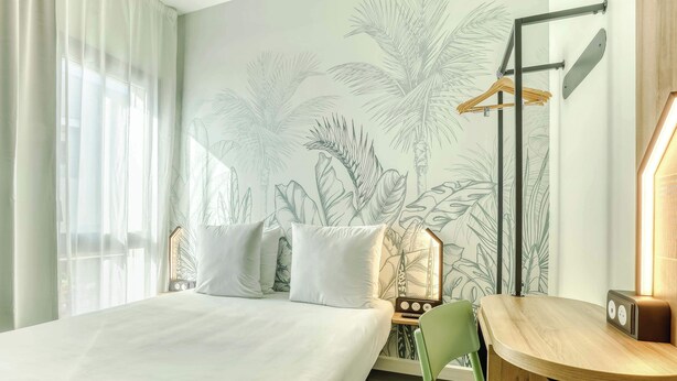 A hotel room with a bed, a desk, a chair, a radio, and a wall with a tropical plant mural. - B&B Hotel La Réunion Aéroport (Sainte Marie)