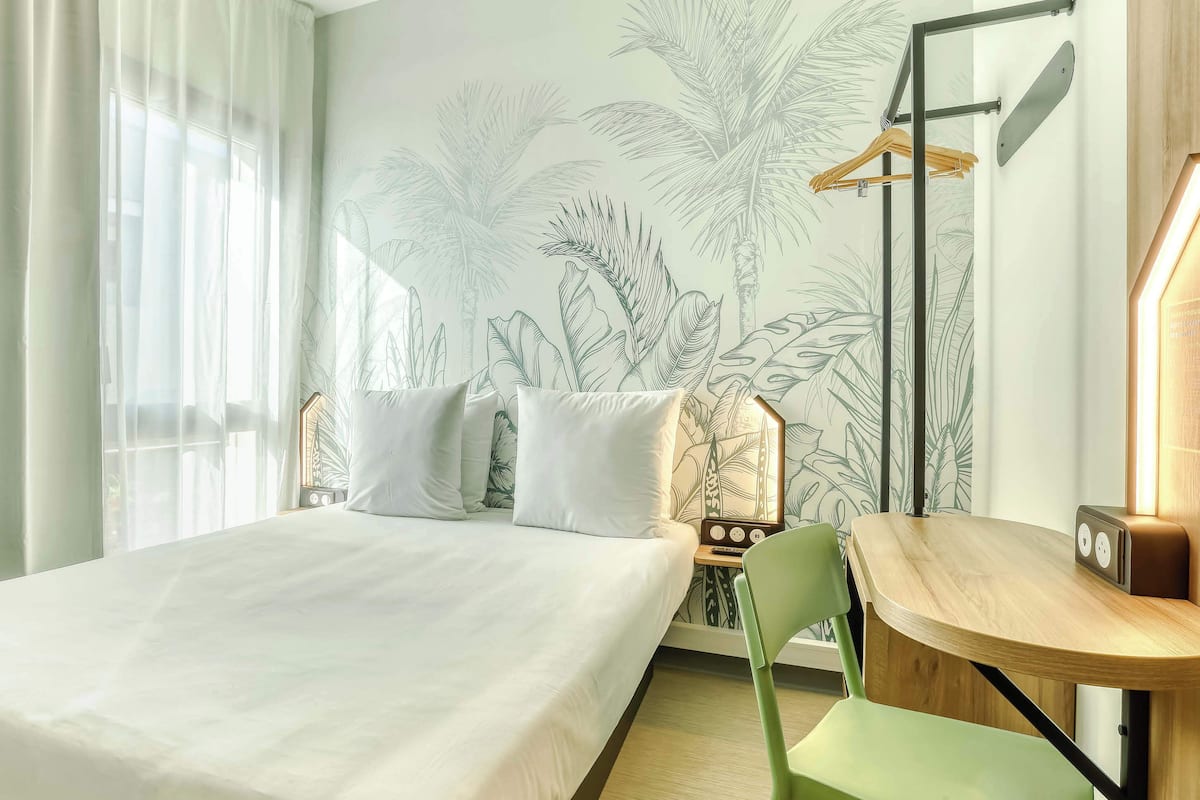 A hotel room with a bed, a desk, a chair, a radio, and a wall with a tropical plant mural.