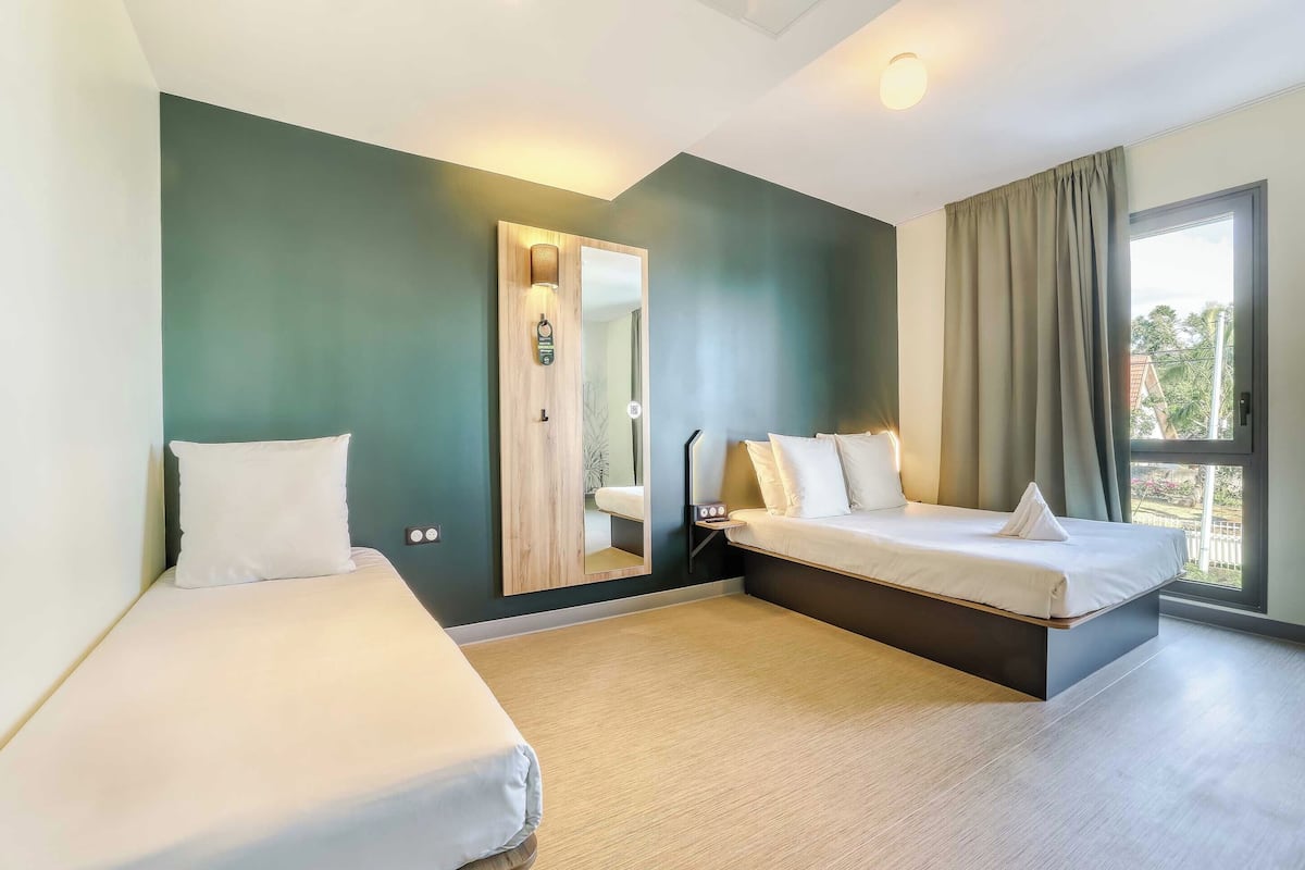 A hotel room with two beds, a large mirror, a small bedside table, and a window with curtains.