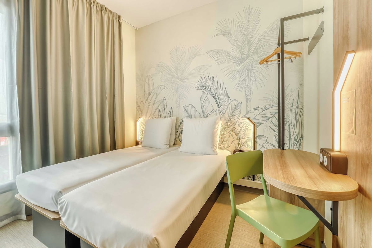 A hotel room with two beds, a green chair, a wooden side table, a mirror, and a wall with a tropical plant mural.