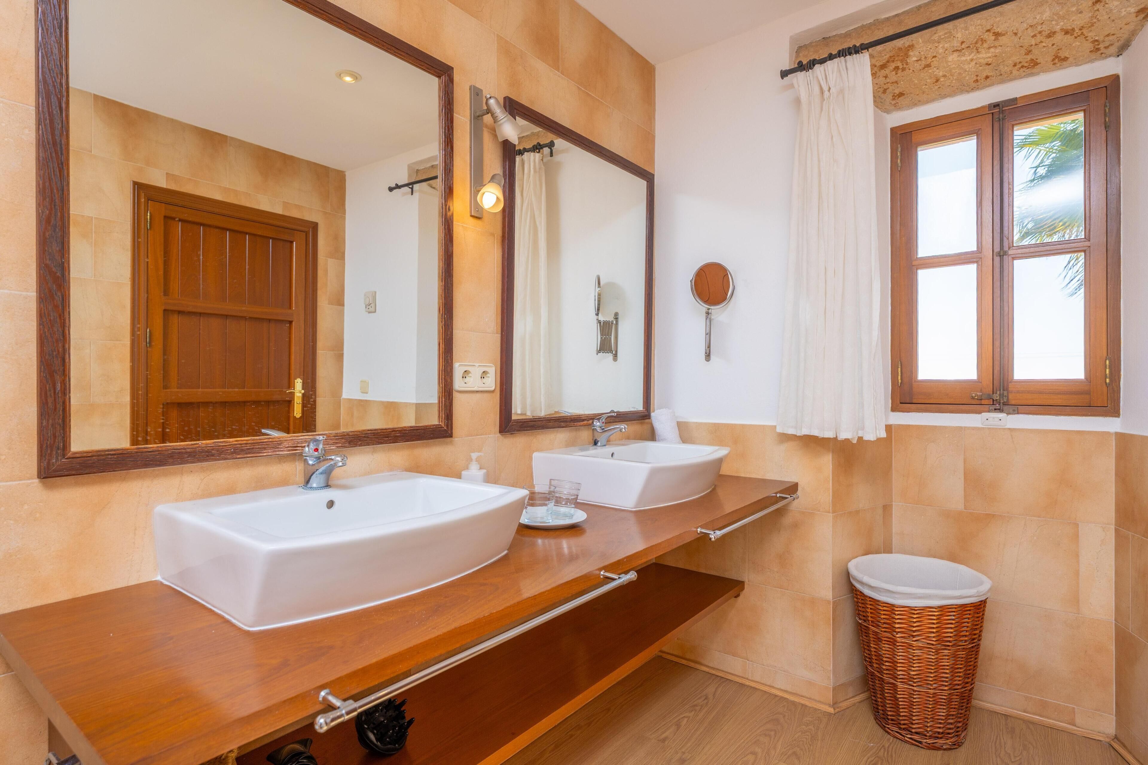 Un baño con dos lavamanos, un espejo grande, una puerta de madera, una ventana con vistas y un cubo de mimbre.