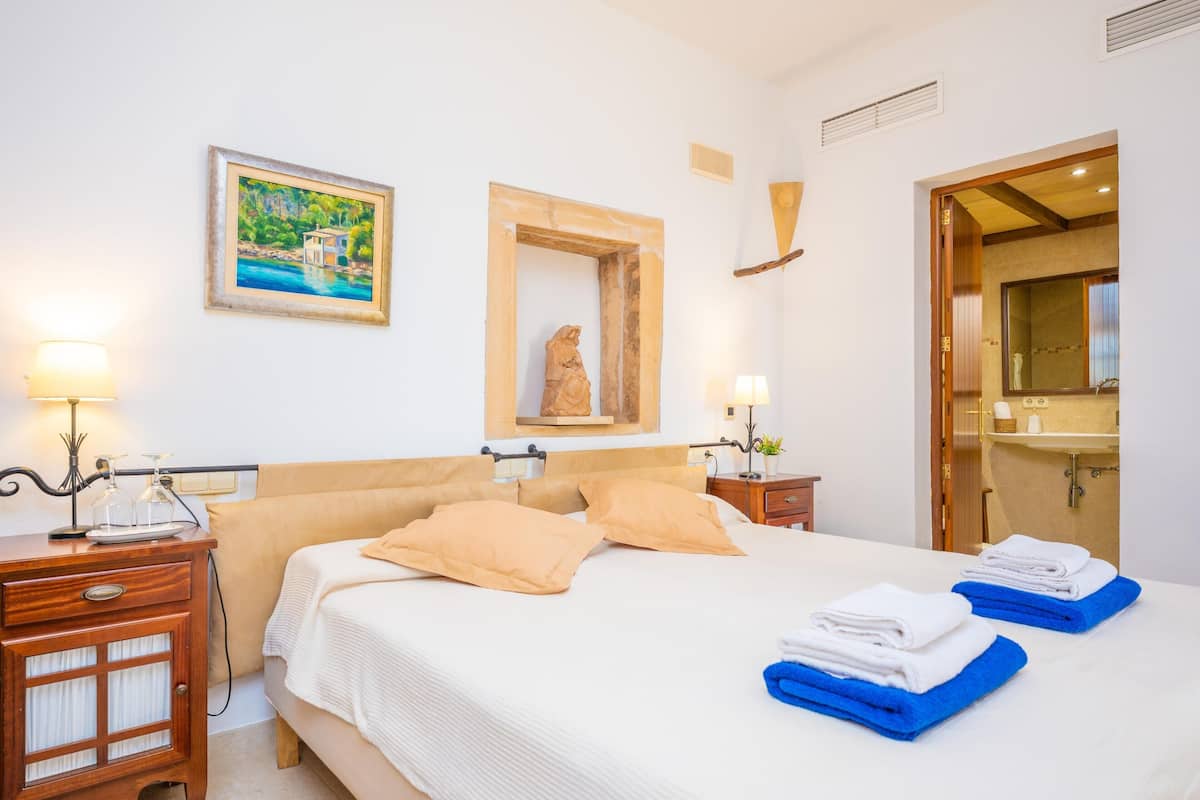 Habitación de hotel con cama, dos mesitas de noche, un tocador, un cuadro, un espejo y un baño visible a través de una puerta abierta.