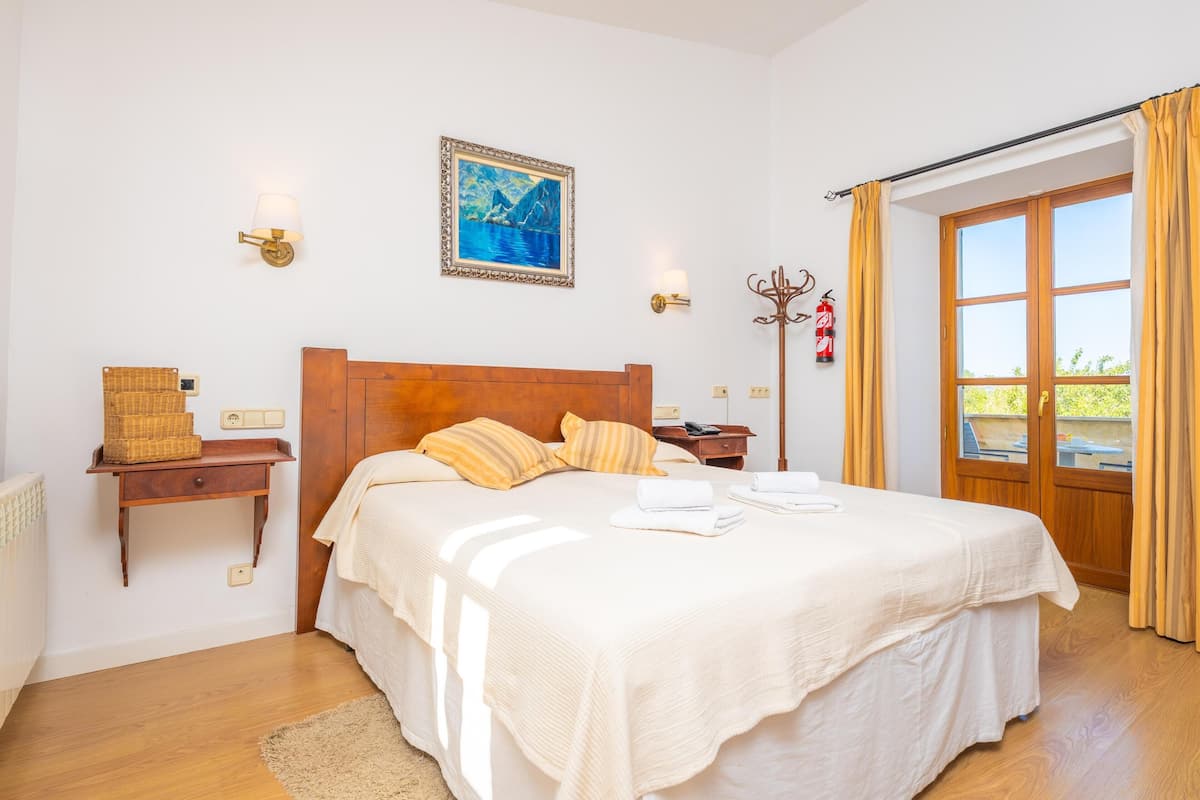 Un dormitorio con cabecera de madera, una cama grande con ropa blanca, una mesita de noche con lámpara, un cuadro enmarcado en la pared y una ventana con cortinas.
