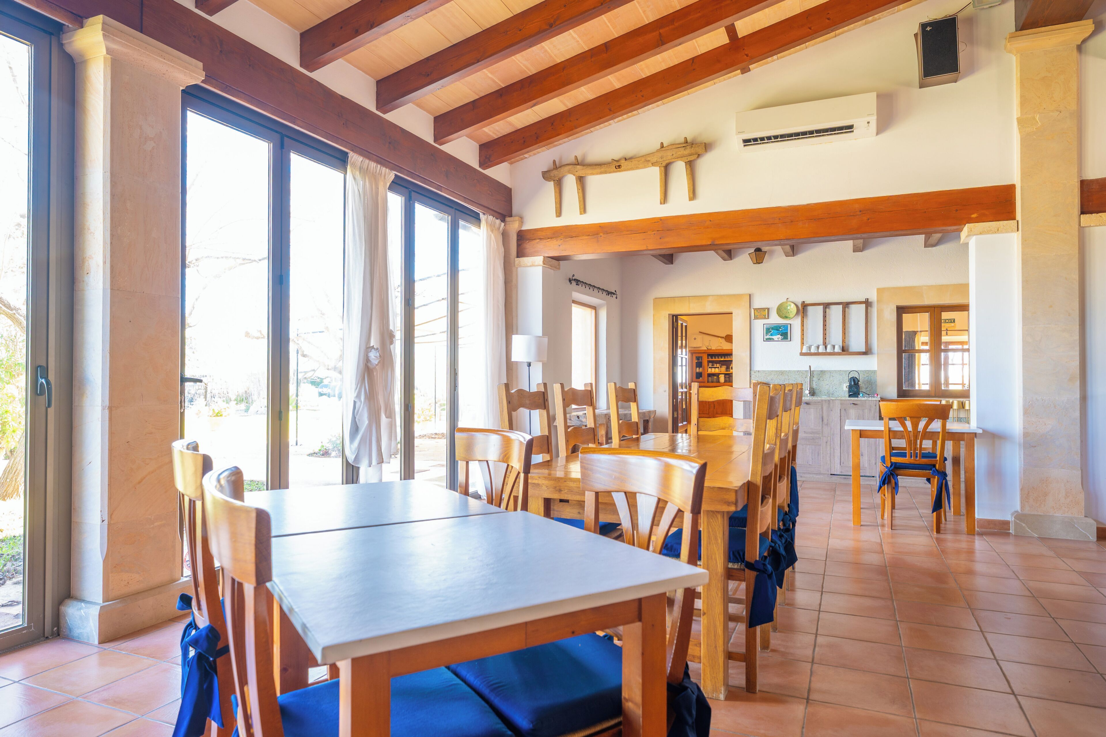 A dining area with wooden tables and chairs, large windows, and a kitchen in the background.