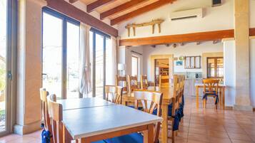 A dining area with wooden tables and chairs, large windows, and a kitchen in the background.