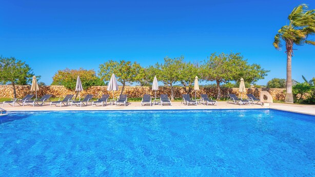 Ein klares, blaues Schwimmbecken mit Liegestühlen und Sonnenschirmen, umgeben von Steinmauern und Bäumen.