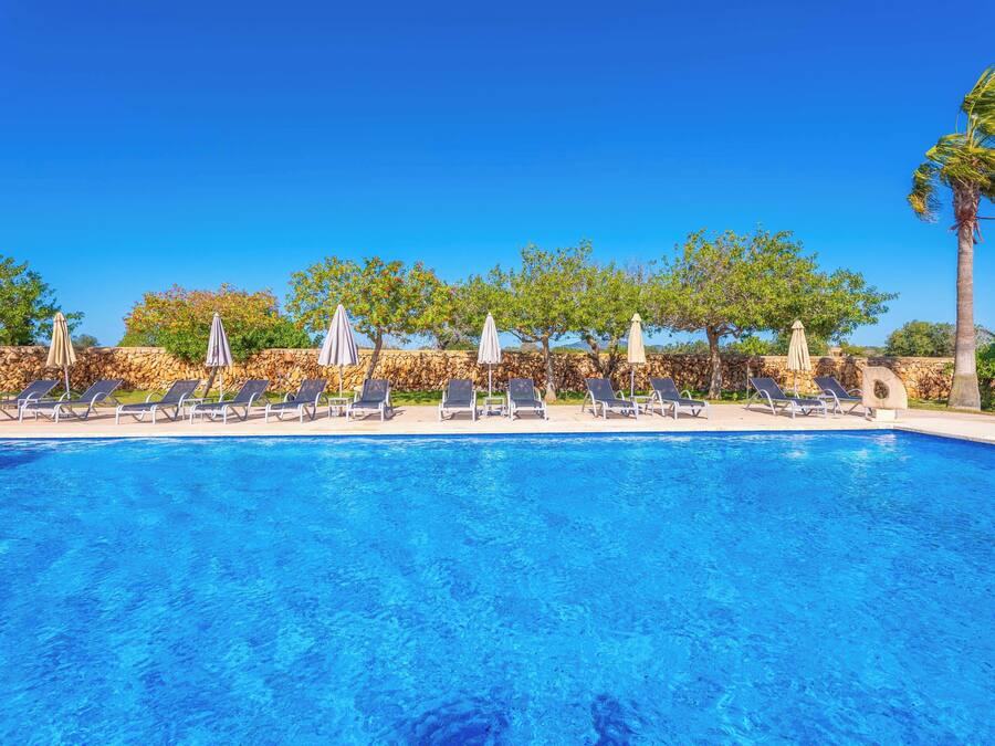 Una piscina azul cristalina con sillas de descanso y sombrillas, rodeada de muros de piedra y árboles.