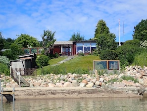 A lakeside property with a wooden dock, a red building with large windows, and a stone pathway leading to the water. - fewo1846 - StrandNest - House with view of the fjord, fireplace, sauna and 3 bed (Glücksburg)