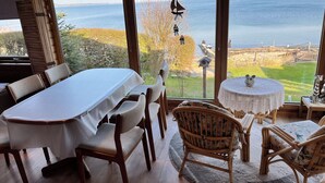 Un espace repas comprenant une table et des chaises, une table ronde agrémentée d’un objet décoratif, et une vue sur la mer grâce à de grandes fenêtres.