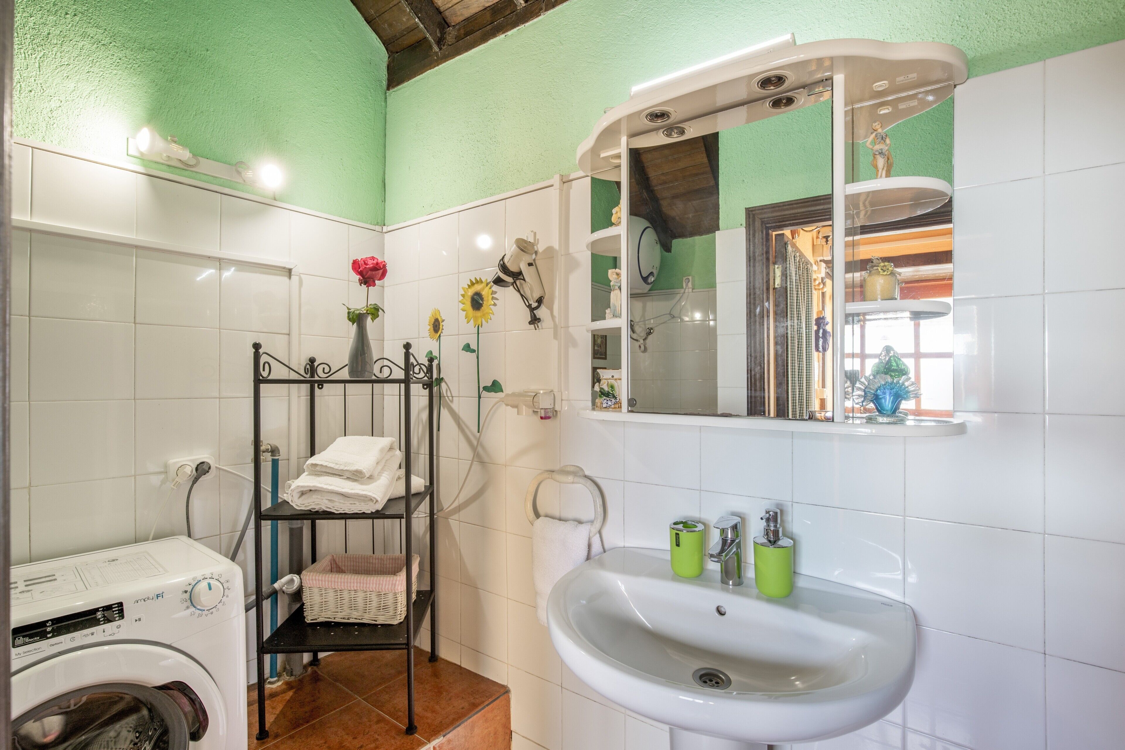 Un baño con lavadora, lavamanos, espejo y estante con toallas y un florero.