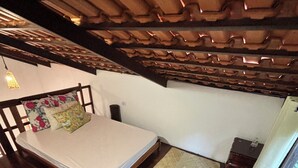 A bedroom with a wooden ceiling, a bed with floral pillows, a small bedside table, and a nightstand. - Pousada Jardim da Vida (Caeté-Açu)