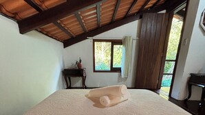 A bedroom with a large bed, a wooden ceiling, a window with curtains, and a small table with a plant. - Pousada Jardim da Vida (Caeté-Açu)