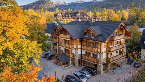 Un lodge a più piani con facciata in legno, un'area di parcheggio e montagne sullo sfondo.