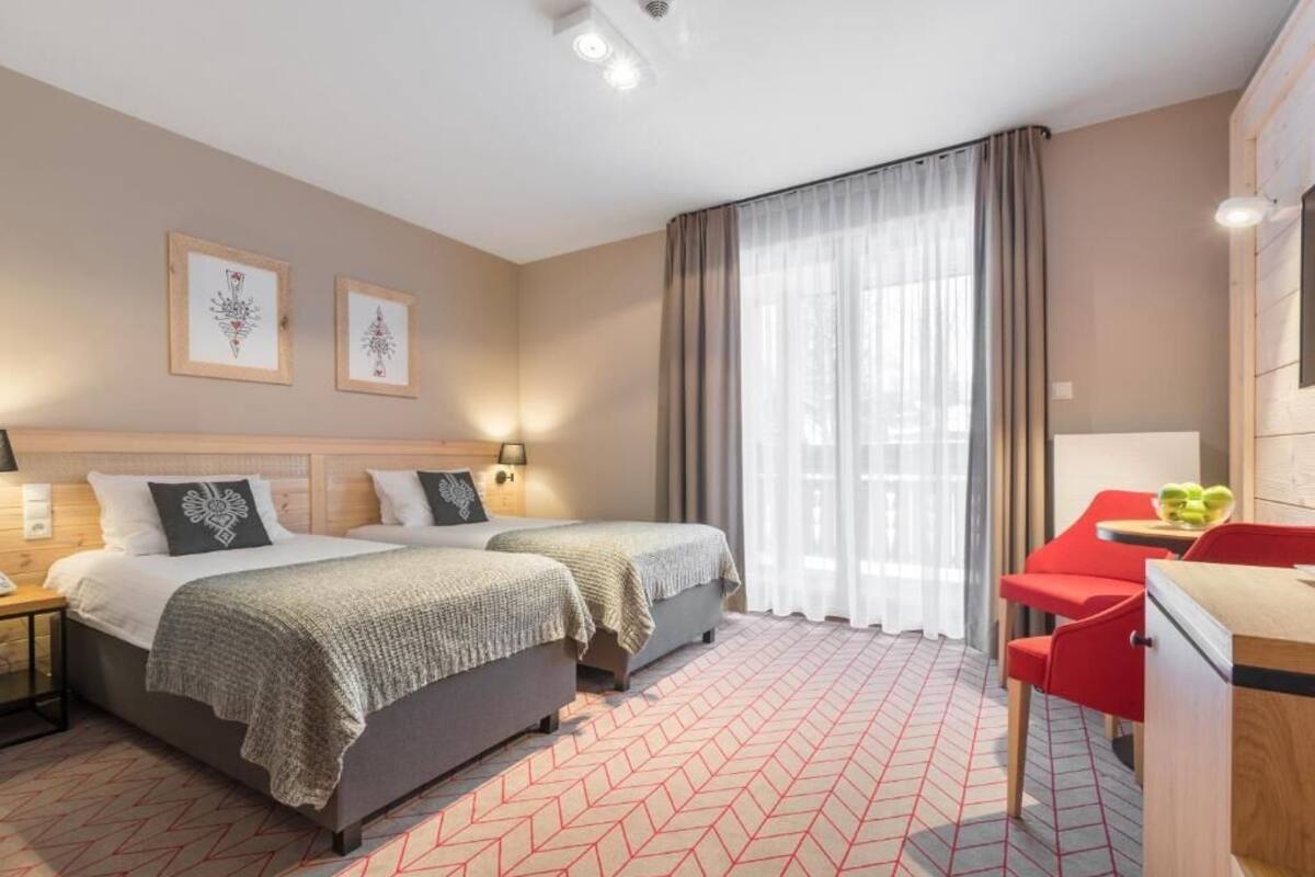 Ein Hotelzimmer mit zwei Betten, einem Schreibtisch mit Stuhl, einem Fenster mit Vorhängen und Wandbildern.