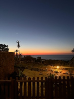 A sunset view from a balcony with a stone wall, wooden fence, and a pathway leading to the ocean. - Poseida Bay - Beautiful Sea View (Paduledda)