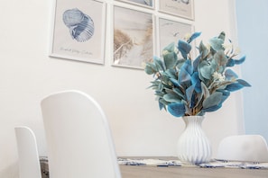 A white chair, a white vase with blue foliage, and framed artwork on a wall. - Poseida Bay - Beautiful Sea View (Paduledda)