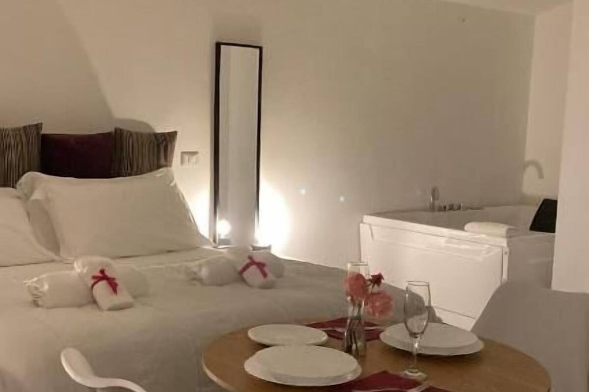 A hotel room with a bed, a bedside table with a lamp, a round dining table with chairs, and a large mirror on the wall.