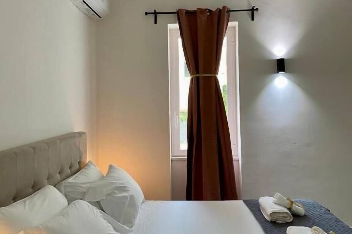 A hotel room with a bed, a window with curtains, an air conditioning unit, and a wall-mounted light fixture.