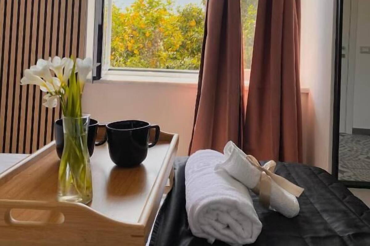 A room with a bed, a bedside table with a tray, a vase with flowers, and a window with curtains.