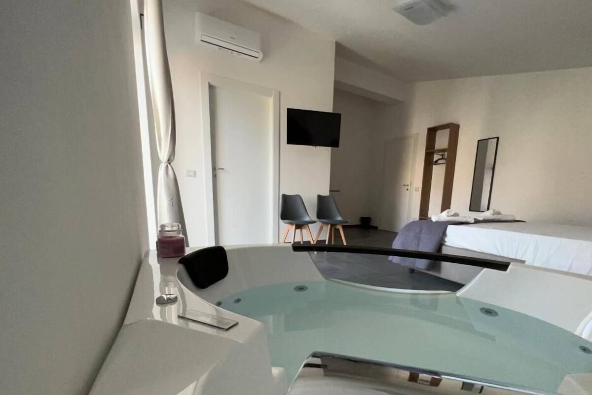 A modern hotel room with a glass-enclosed jacuzzi, a flat-screen TV mounted on the wall, a bed with white linens, and a small seating area with two chairs.