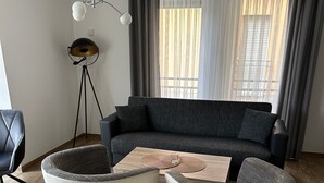 Ett modernt vardagsrum med en mörkgrå soffa, två vita fåtöljer, ett träbord, en golvlampa och ett stort fönster med gardiner.