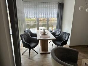 A dining area with a table and four chairs, a wicker chair, and a view of a body of water through large windows. - Vacation apartment with a view of the Rauenstein bastion in Saxon Switzerland (Stadt Wehlen)