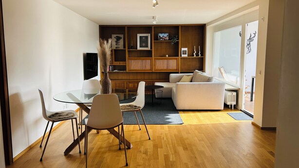 Een moderne woonkamer met een glazen eettafel, witte stoelen, een bank en een houten boekenkast.