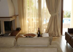 A living room with a sofa, a table with a bottle and remote control, and a view of a pool through sheer curtains. - Verde Luxury Villas Skiathos (Troulos)
