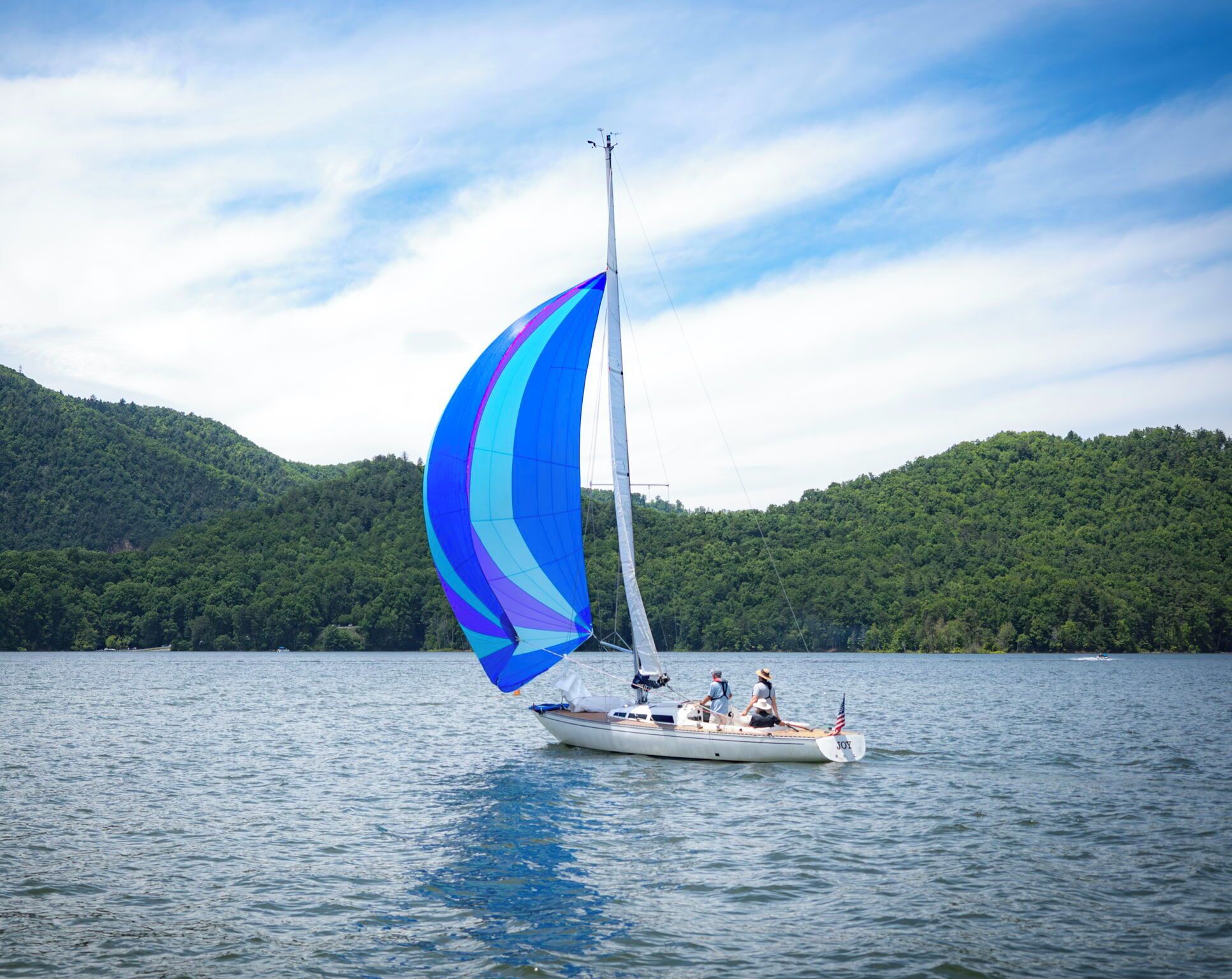 A sailboat with a colorful sail on a calm lake, surrounded by lush green hills.