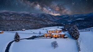 Een besneeuwd landschap met een huis, verlicht in de schemering, omringd door bergen en een sterrenhemel.