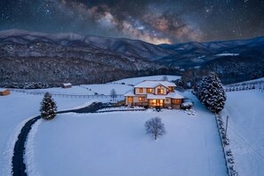 Schneebedeckte Landschaft mit einem Haus, das im Abenddämmerung beleuchtet ist, umgeben von Bergen und einem Sternenhimmel.